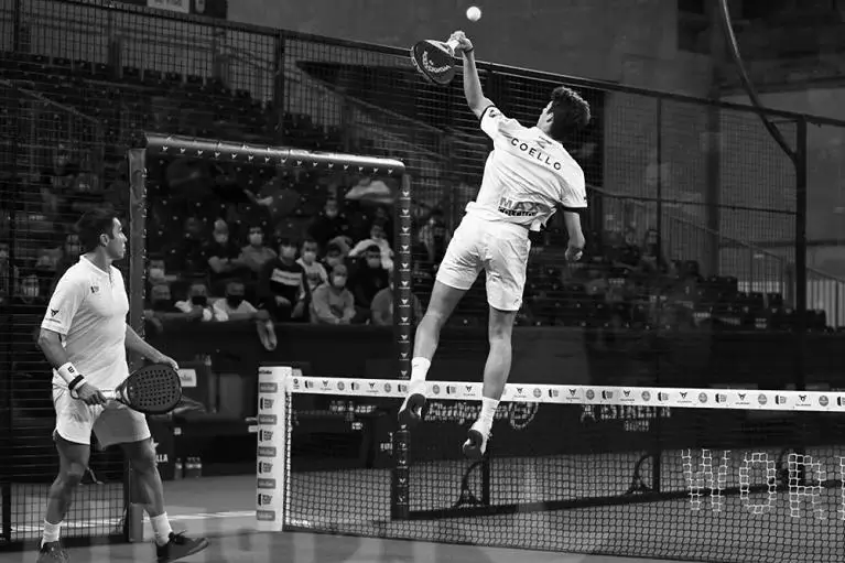 Arturo Coello leaps into the air for a powerful overhead smash, while his partner stands ready at the net during a padel match.