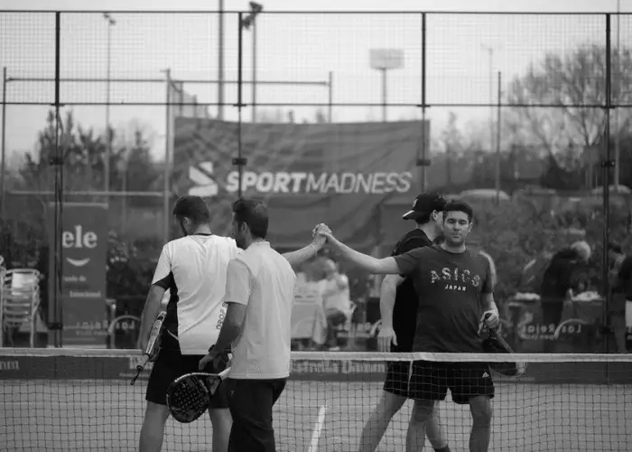 Four padel players meeting at the net for a post-match handshake, with a "Sport Madness" banner in the background.