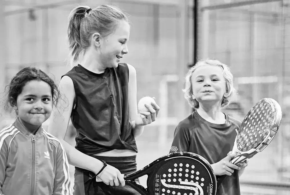 Three young kids happily hold their padel rackets, ready to play, showcasing the joy of starting the sport at an early age.