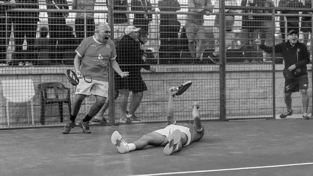 Two older padel players celebrating a victory, one standing and cheering while the other lies on the court in joy.