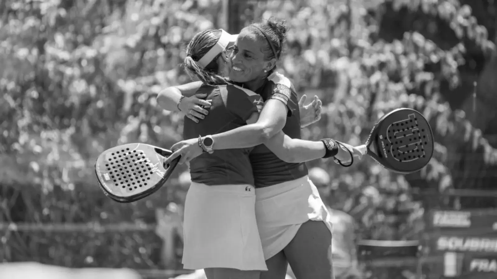 Two female padel players in a joyful embrace on the court after a match, holding their paddles.