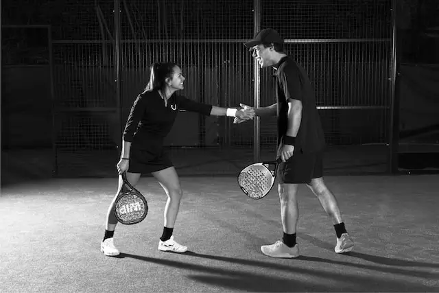 Two padel players shaking hands on the court after a match, both holding rackets and smiling under court lights.